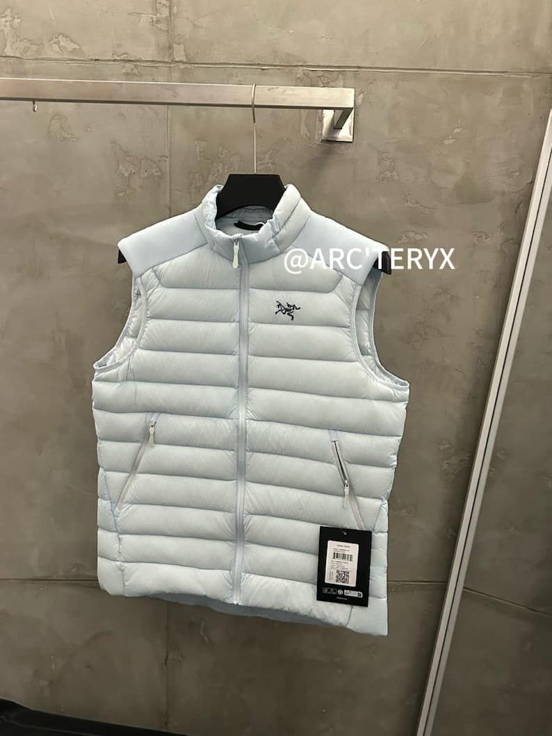 May include: A light blue puffer vest with a high collar and a zipper closure. The vest has two side pockets and a small logo on the chest. The text "@ARCTERYX" is visible. A tag with a barcode hangs from the vest.