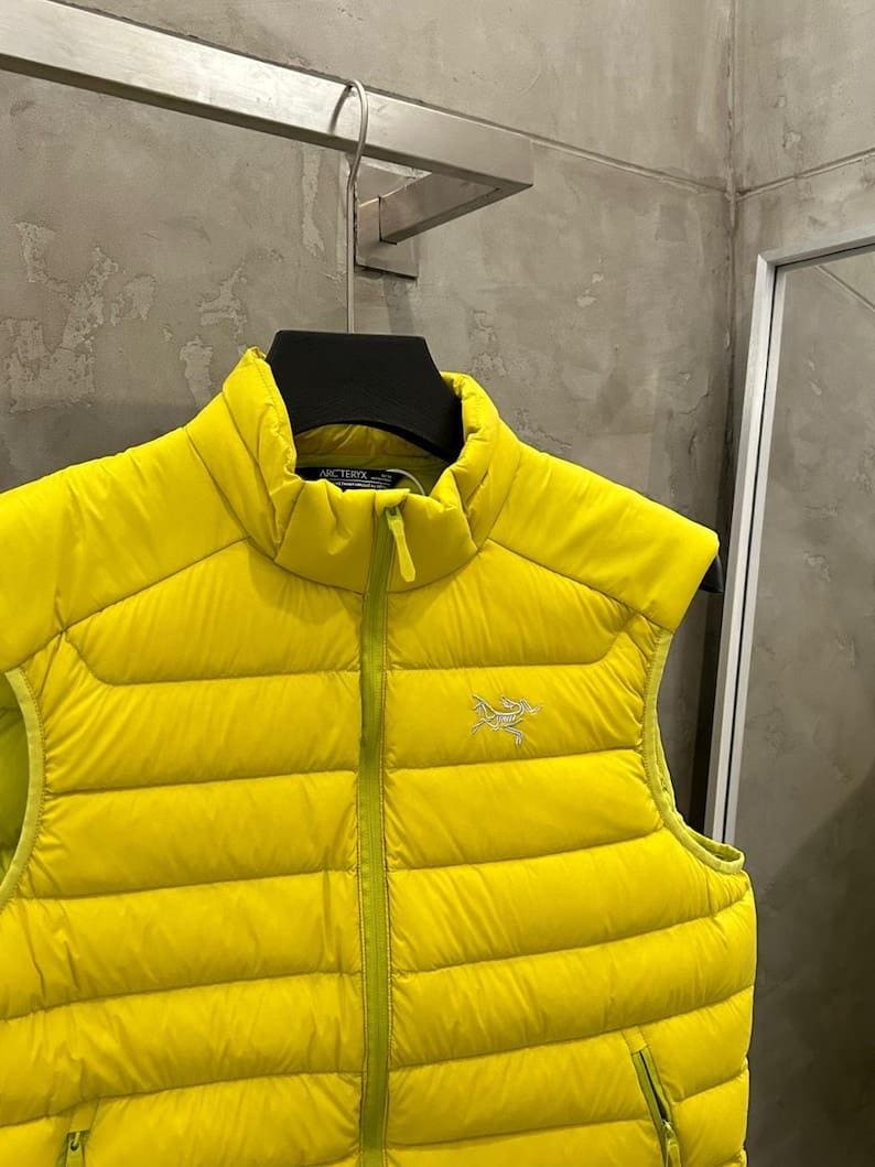 May include: A bright yellow puffer vest with a high collar and a front zipper. The vest has a quilted design and a small logo on the chest. The vest is sleeveless and appears to be made of a lightweight material.