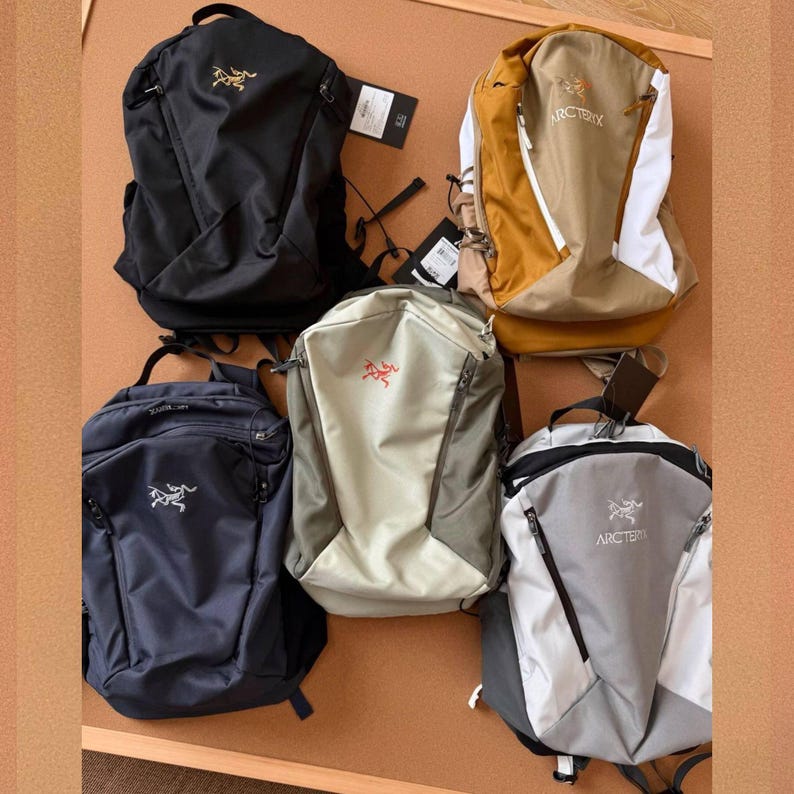 May include: Five backpacks in various colors, including black, navy blue, tan, and gray. Each backpack features a logo and multiple compartments. The backpacks are displayed on a brown surface.