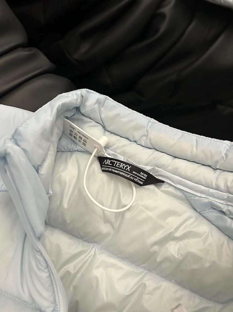 May include: Light blue quilted jacket with a tag that reads "ARC'TERYX". The jacket is made of a smooth, padded material. The tag also includes the size M/M and the text "MADE IN VIETNAM/FABRIQU&Eacute; AU VIETNAM".
