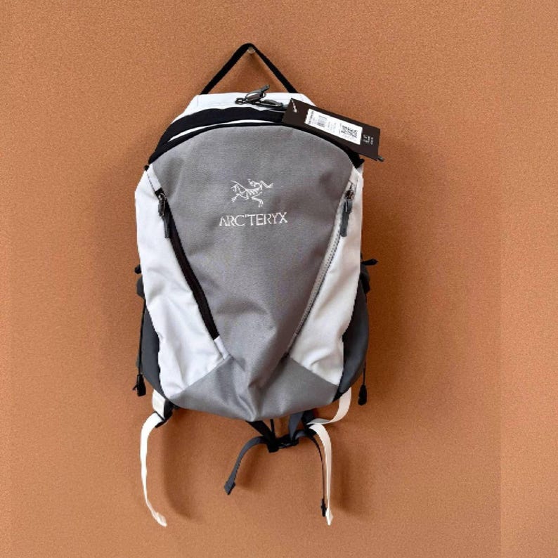 May include: A gray, white, and black backpack with the brand name "ARC'TERYX" printed on the front. The backpack has a black handle, a zipper, and white straps. A black tag hangs from the top.