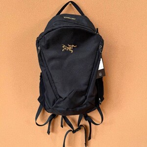 May include: Black backpack with a top handle and adjustable straps. The backpack features a gold logo and the brand name in gold lettering. A tag hangs from the side. The backpack is hanging against a tan background.