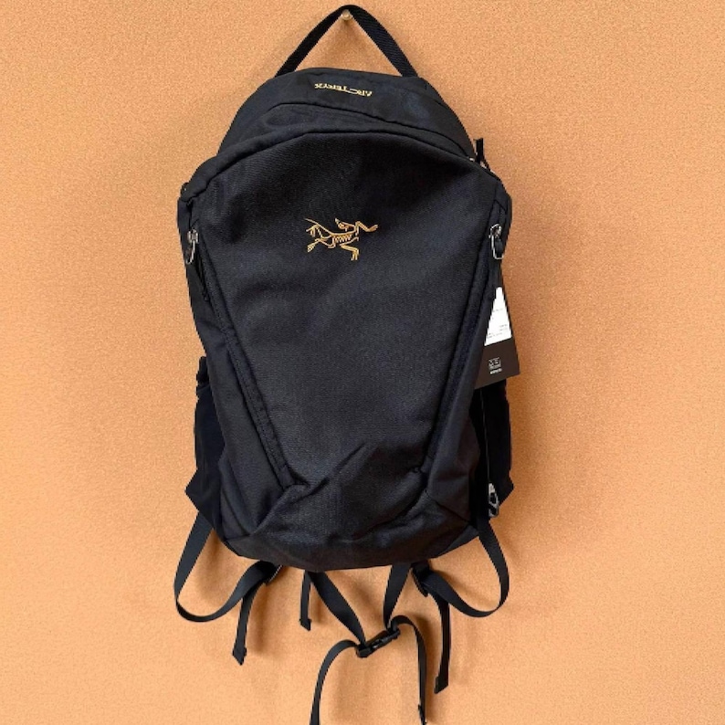 May include: Black backpack with a top handle and adjustable straps. The backpack features a gold logo and the brand name in gold lettering. A tag hangs from the side. The backpack is hanging against a tan background.
