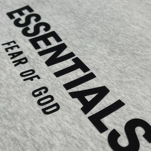 Puede incluir: Primer plano de una camiseta gris claro con la palabra "ESSENTIALS" impresa en letras negras grandes y en negrita. Debajo, el texto "FEAR OF GOD" est&aacute; impreso en una fuente m&aacute;s peque&ntilde;a.
