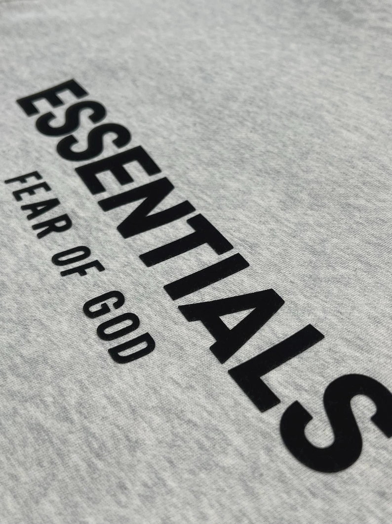 Puede incluir: Primer plano de una camiseta gris claro con la palabra "ESSENTIALS" impresa en letras negras grandes y en negrita. Debajo, el texto "FEAR OF GOD" est&aacute; impreso en una fuente m&aacute;s peque&ntilde;a.