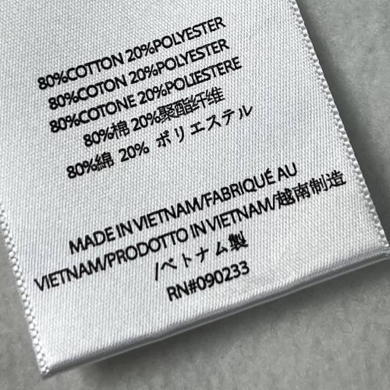Puede incluir: Primer plano de una etiqueta de tela blanca con texto negro. La etiqueta indica la composici&oacute;n de la tela: 80% algod&oacute;n y 20% poli&eacute;ster en varios idiomas. Tambi&eacute;n indica 'Hecho en Vietnam'. RN #090233.