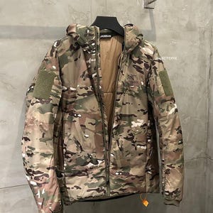 Op de afbeelding: Een camouflage jack met capuchon en een lange ritssluiting. De jas heeft een mix van groene, bruine en bruinachtige kleuren. De merknaam "Arc'teryx" is zichtbaar op de borst. De jas heeft een patch op de linkermouw.