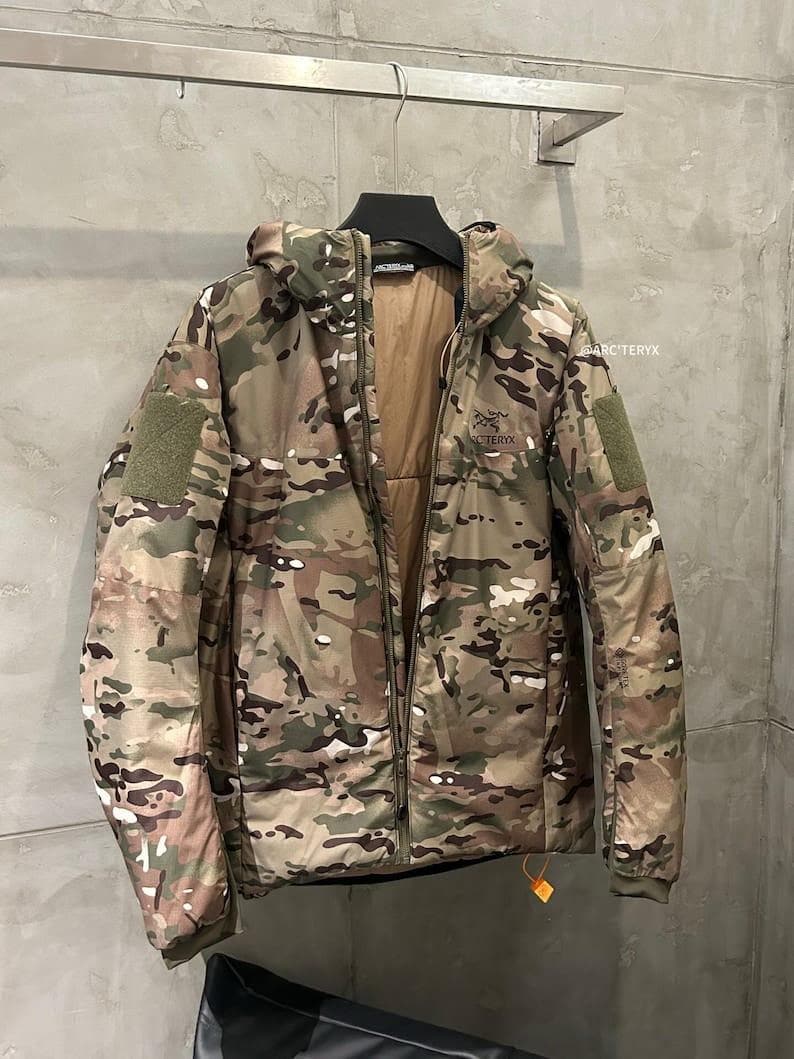 Op de afbeelding: Een camouflage jack met capuchon en een lange ritssluiting. De jas heeft een mix van groene, bruine en bruinachtige kleuren. De merknaam "Arc'teryx" is zichtbaar op de borst. De jas heeft een patch op de linkermouw.