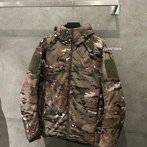 Op de afbeelding: Een camouflagepatroon jas met een capuchon, ritssluiting en twee groene patches op de mouwen. De jas, met het Arc'teryx logo, hangt aan een metalen stang tegen een grijze muur.