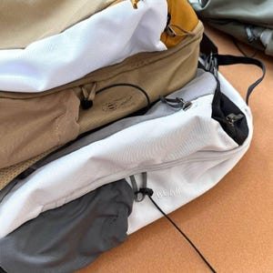 May include: A multi-toned backpack with a tan, white, and gray color scheme. The bag features multiple compartments, zippers, and a black strap. The word "BEAMS" is visible on the side of the bag.
