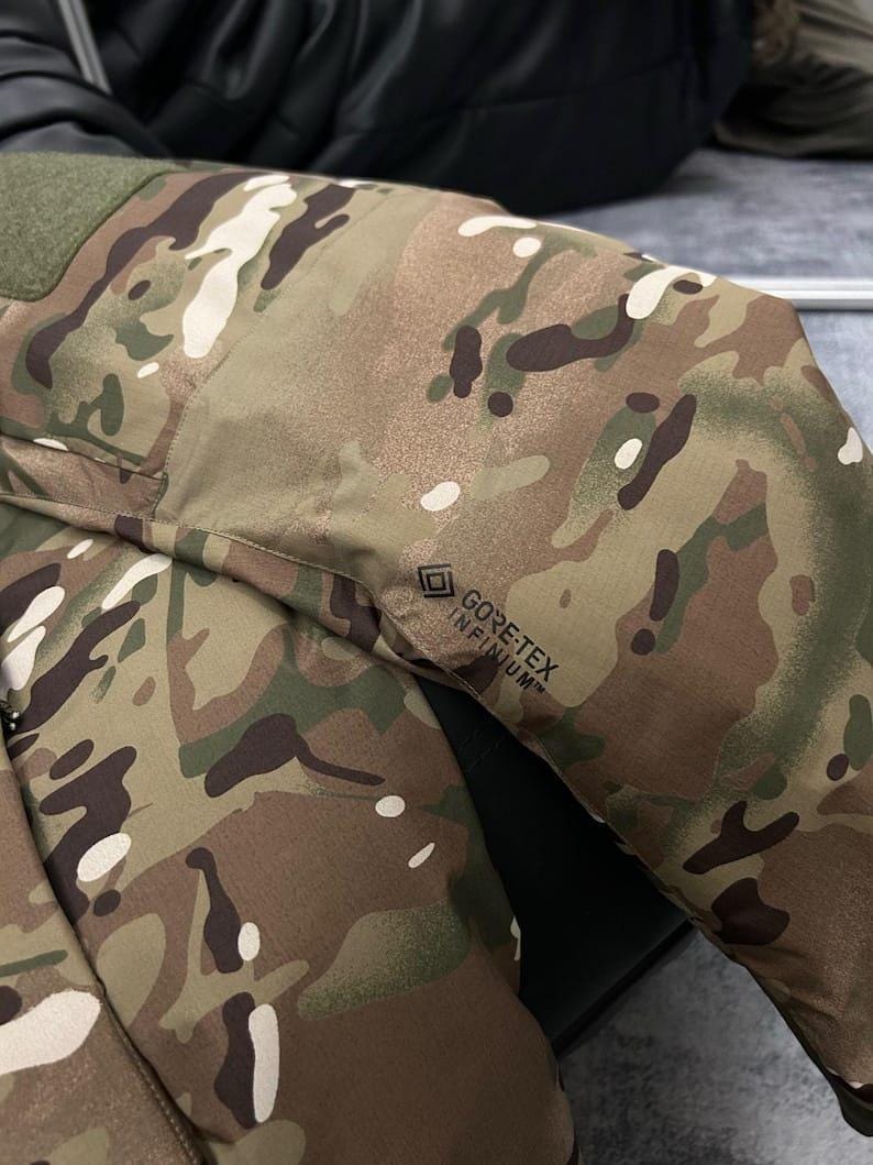 Op de afbeelding: Close-up van een camouflagejas met groene, bruine en witte patronen. De jas heeft een groene patch op de schouder en het "GORE-TEX INFINIUM" logo. De stof lijkt duurzaam en weerbestendig.