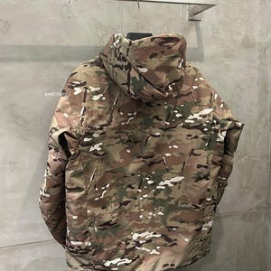 Op de afbeelding: Een camouflagejas met capuchon, met een patroon van bruin, groen en wit. De jas hangt aan een metalen rek tegen een grijze muur. De merknaam "@ARCTERYX" is zichtbaar op de jas.