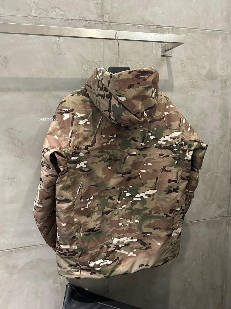 Op de afbeelding: Een camouflagejas met capuchon, met een patroon van bruin, groen en wit. De jas hangt aan een metalen rek tegen een grijze muur. De merknaam "@ARCTERYX" is zichtbaar op de jas.