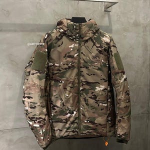 Op de afbeelding: Een camouflagejas met capuchon, ritssluiting en een klein logo op de borst. De jas heeft een groene patch op de linkermouw en hangt aan een metalen stang. Het patroon van de jas bevat tinten groen, bruin en wit.