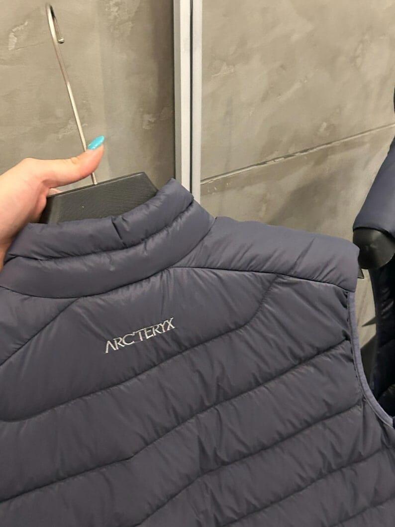 May include: A dark gray quilted vest with the brand name "ARC'TERYX" printed in white. The vest is hanging on a black hanger. The vest has a high collar and a diagonal quilted pattern.