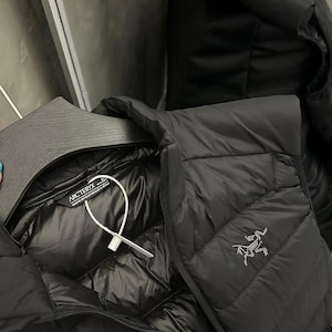 May include: Black quilted down jacket with a shiny interior. The jacket features a zipper closure and a small logo on the chest. The jacket is hanging on a black hanger.