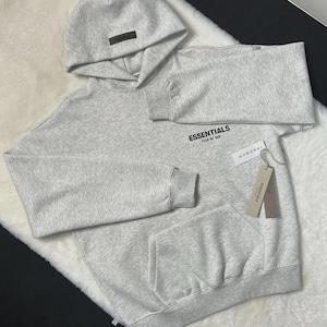 Puede incluir: Una sudadera con capucha gris claro con capucha y bolsillo delantero. La palabra "ESSENTIALS" est&aacute; impresa en el pecho. Una etiqueta que dice "PACSUN" est&aacute; adjunta a la sudadera con capucha.