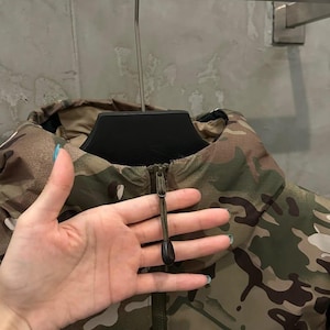 Op de afbeelding: Camouflage jas met een hoge kraag en ritssluiting. De jas heeft een bruin, groen en beige kleurenschema. Het Arc'teryx logo is zichtbaar aan de rechter onderkant.