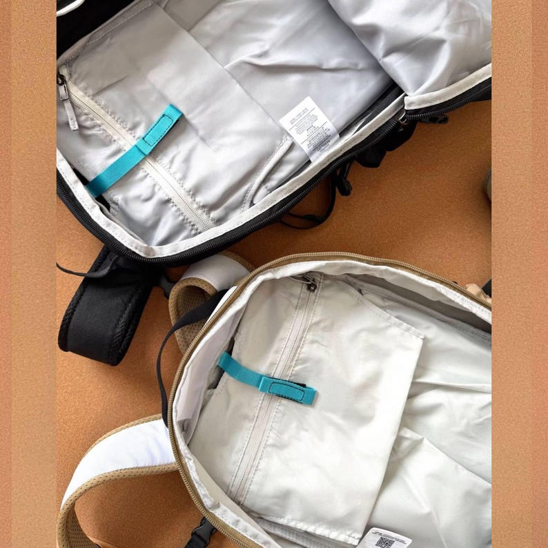 May include: Two open backpacks, one black and one tan, showcasing their interior compartments. The black backpack has a gray lining with a teal strap, while the tan one features a similar design. Both have zippered pockets and padded straps.