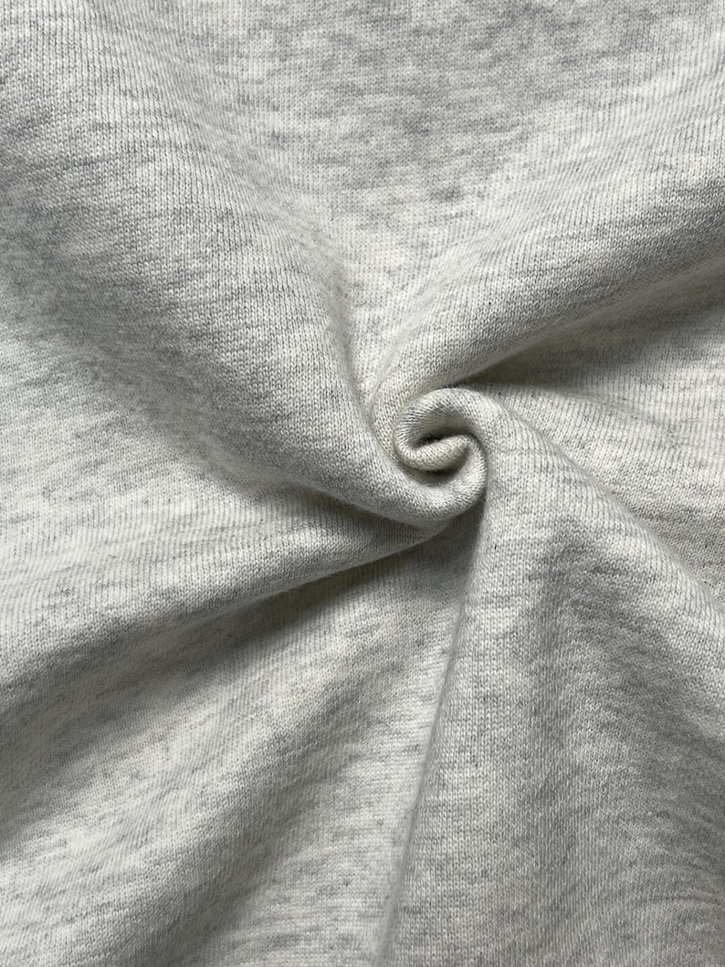 Puede incluir: Primer plano de una tela gris claro con un sutil patr&oacute;n moteado. La tela es suave y parece estar tejida, con textura y pliegues visibles. El material est&aacute; tejido apretado, creando una superficie lisa.