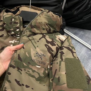 Op de afbeelding: Camouflage jas in groen, bruin en beige, met het Arc'teryx logo. De jas heeft een ritssluiting en een capuchon. Een groene patch is zichtbaar op de mouw.