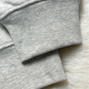 Puede incluir: Primer plano del pu&ntilde;o de una sudadera gris claro. El pu&ntilde;o es acanalado y tiene una costura visible. La tela parece ser un material suave y tejido. El fondo es una superficie blanca texturizada y borrosa.