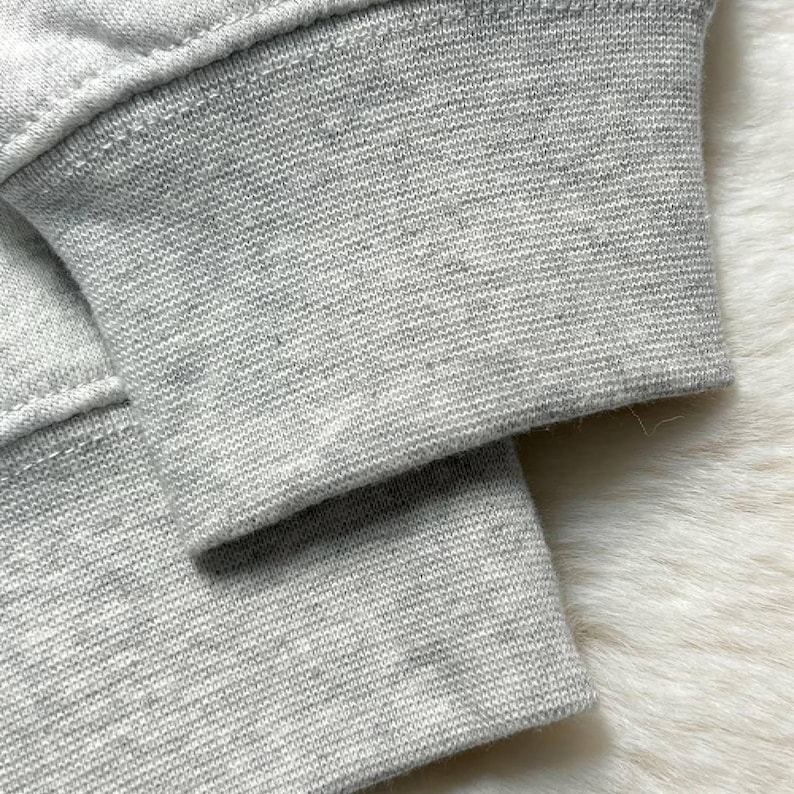 Puede incluir: Primer plano del pu&ntilde;o de una sudadera gris claro. El pu&ntilde;o es acanalado y tiene una costura visible. La tela parece ser un material suave y tejido. El fondo es una superficie blanca texturizada y borrosa.