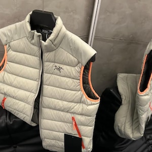 May include: A light gray puffer vest with orange trim and zippers. The vest has a black interior and a small logo on the chest. The vest is hanging on a black hanger.