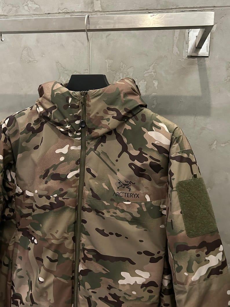Op de afbeelding: Camouflagepatroon jas met een capuchon en ritssluiting. De jas heeft het "ARC'TERYX" logo op de borst en een klittenband patch op de mouw. Het kleurenpalet bevat tinten groen, bruin en beige.