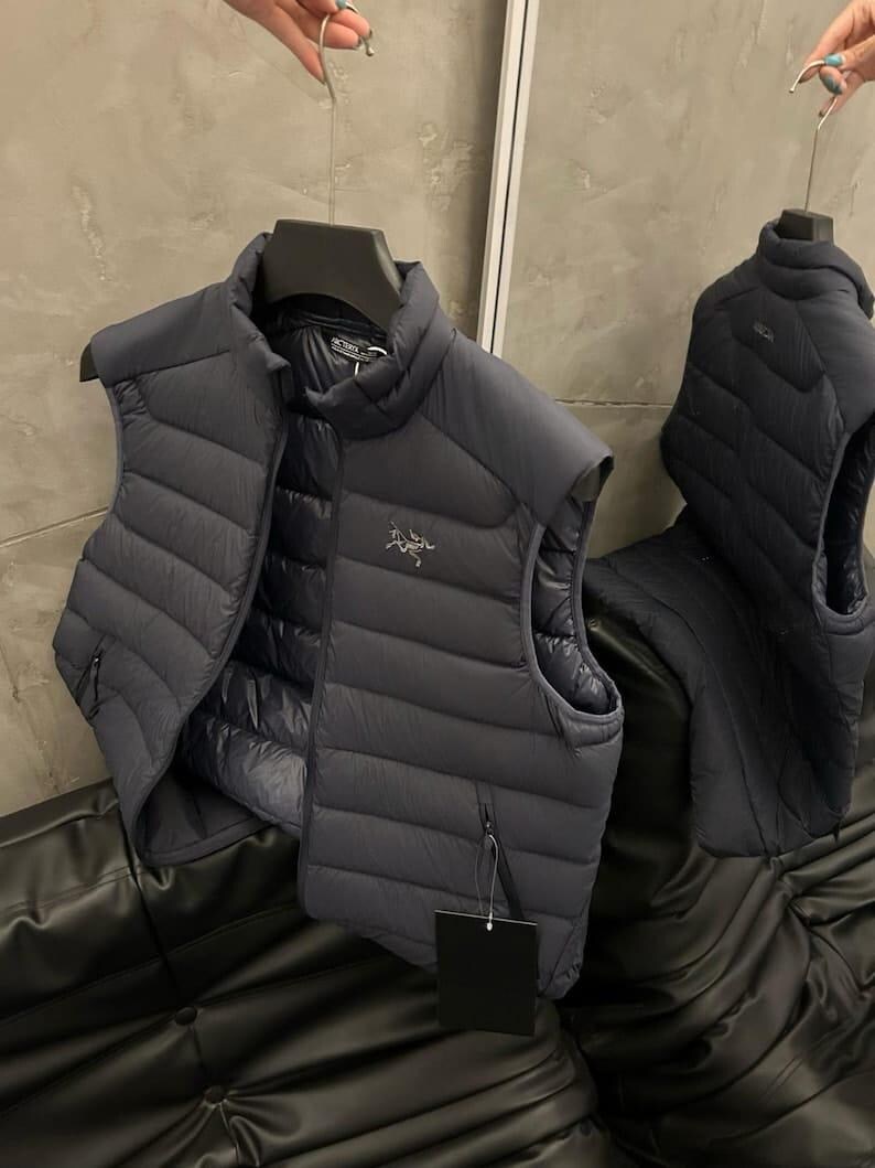 May include: Two dark gray puffer vests hanging on hangers. The vests have a quilted design and a small logo on the chest. One vest is open, revealing a black interior. A black tag hangs from one of the vests.