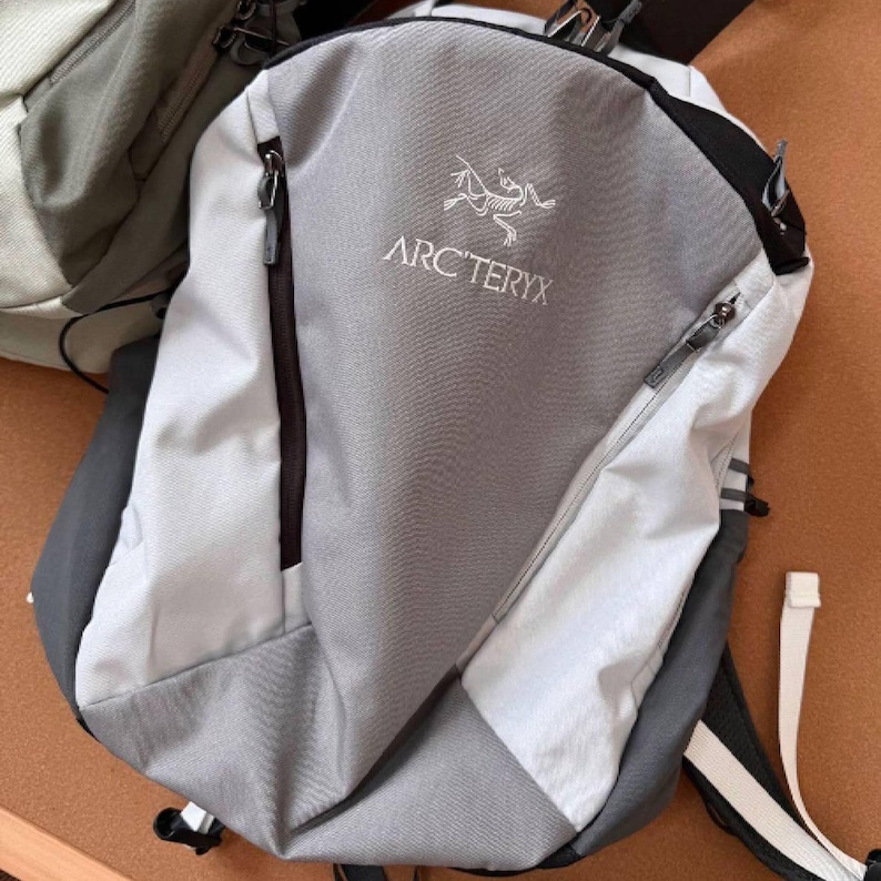 May include: A gray and white backpack with the Arc'teryx logo and text. The backpack has a zippered front pocket and a black and white strap. The backpack is made of a durable material.
