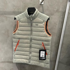 May include: A light gray puffer vest with orange trim and zippers. The vest has a high collar and a small logo on the chest. It is hanging on a black hanger against a gray wall.