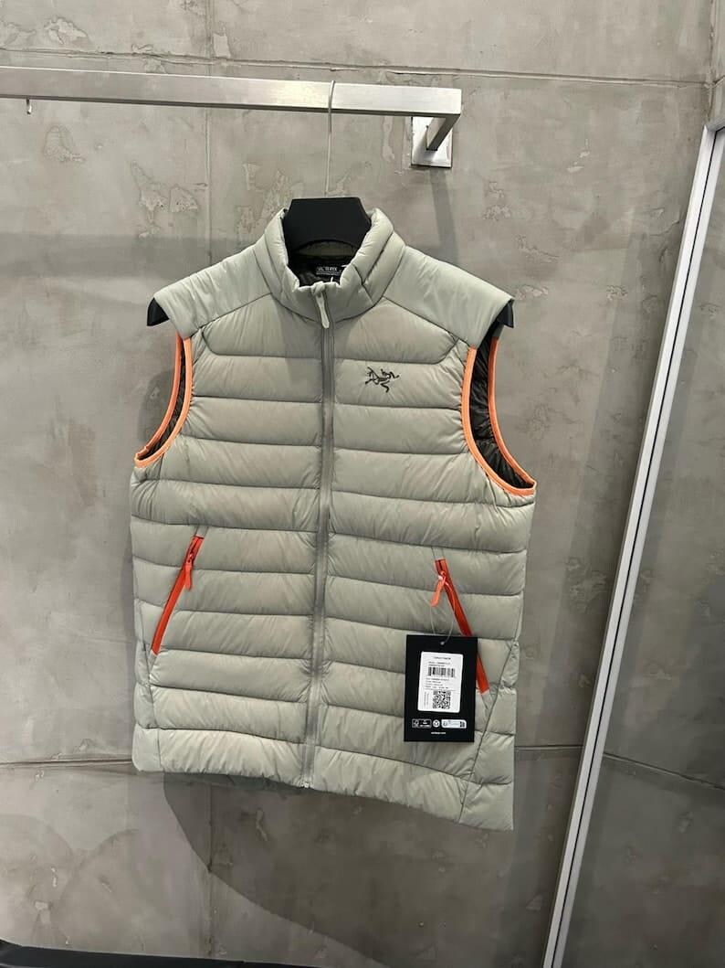 May include: A light gray puffer vest with orange trim and zippers. The vest has a high collar and a small logo on the chest. It is hanging on a black hanger against a gray wall.