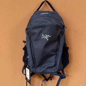 May include: Navy blue backpack with a white logo of a dinosaur-like creature. The backpack has a top handle, multiple zippered compartments, and adjustable straps. A tag hangs from the side. The brand name "ARCTERYX" is visible at the top.