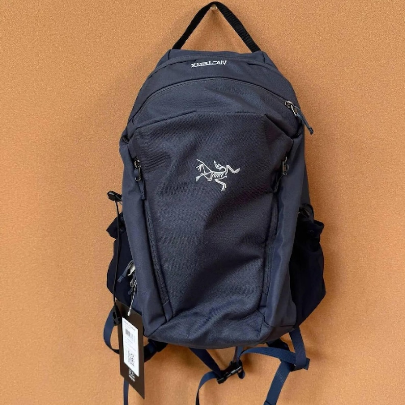 May include: Navy blue backpack with a white logo of a dinosaur-like creature. The backpack has a top handle, multiple zippered compartments, and adjustable straps. A tag hangs from the side. The brand name "ARCTERYX" is visible at the top.