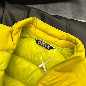 May include: Bright yellow puffer jacket with a quilted design. The jacket features a high collar and a tag with the brand name "Arc'teryx". The jacket is made of a smooth, shiny material.