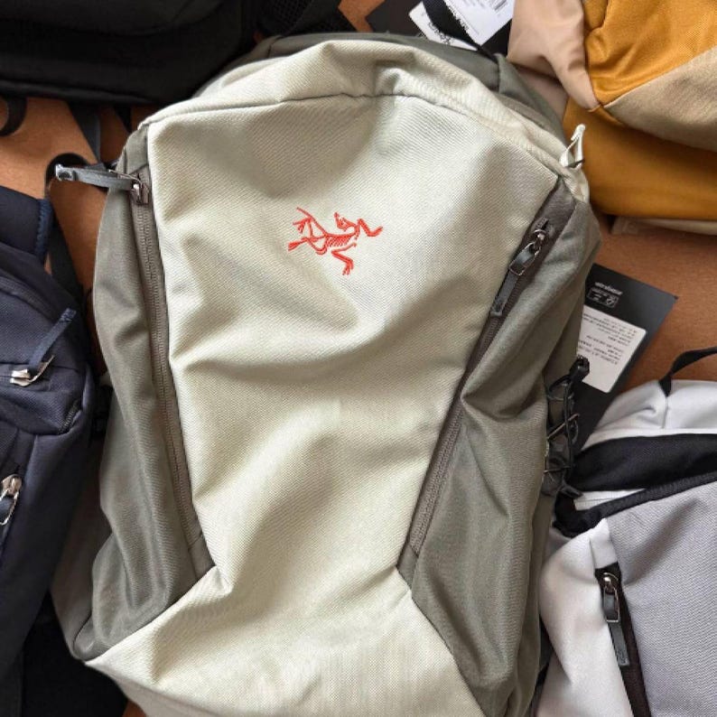 May include: A light green and beige backpack with a red logo. The backpack has multiple zippered compartments and adjustable straps. The bag is surrounded by other bags.