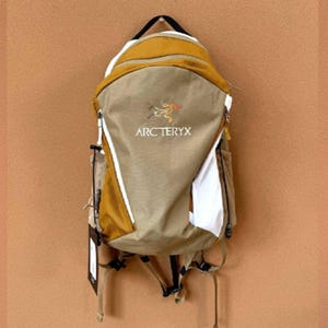 May include: A tan and mustard yellow backpack with the Arc'teryx logo. The backpack has white accents and a black handle. The bag is hanging against a brown background. The backpack has multiple compartments and straps.