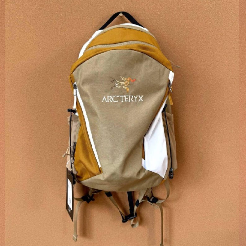 May include: A tan and mustard yellow backpack with the Arc'teryx logo. The backpack has white accents and a black handle. The bag is hanging against a brown background. The backpack has multiple compartments and straps.