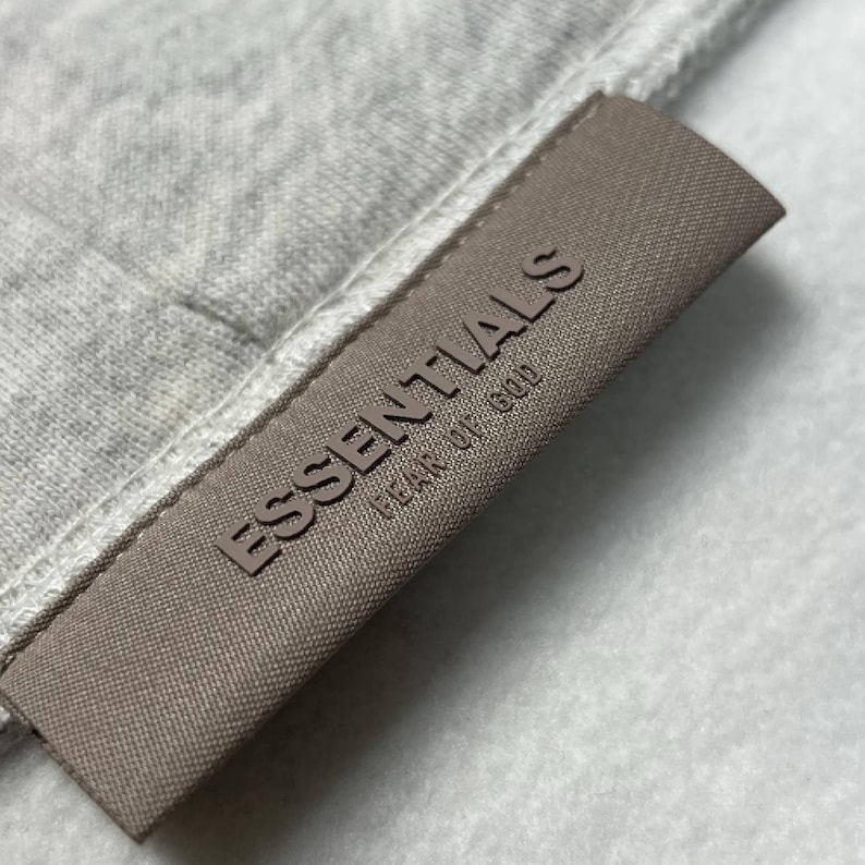 Puede incluir: Primer plano de una tela gris con una etiqueta marr&oacute;n. La etiqueta dice "ESSENTIALS FEAR OF GOD" en relieve. La tela parece ser un material suave, posiblemente una sudadera.