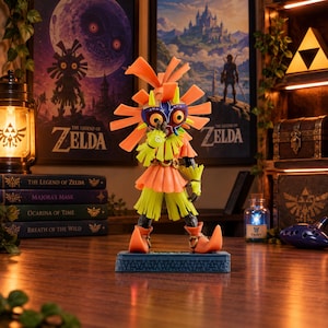 Peut inclure: Une figurine de collection représentant Skull Kid de The Legend of Zelda, avec une tenue orange et jaune vive, le masque de Majora violet et rouge, et une coiffe orange à pointes, posée sur un socle bleu rectangulaire aux motifs sculptés.