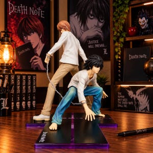 May include: Two anime character figures connected by a silver metal chain. One figure stands wearing a white long-sleeve shirt and tan trousers, while the other crouches wearing a white long-sleeve shirt and blue trousers. They are mounted on a black base.