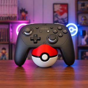 May include: A black Nintendo Switch Pro controller resting on a red and white spherical Pokeball-themed stand. The controller features standard black buttons, a directional pad, two analog sticks, and the Nintendo Switch logo printed in white.