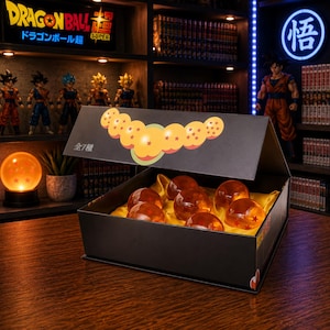 Könnte beinhalten: Eine schwarze rechteckige Präsentationsbox mit sieben durchscheinenden orangefarbenen Kugeln, die jeweils mit roten Sternen markiert sind. Der Deckel zeigt eine Grafik von sieben Kugeln und den japanischen Text "全7種". Das Innere ist mit gelbem Satin ausgekleidet.