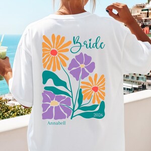 Könnte beinhalten: Ein weißes T-Shirt mit einer bunten Blumengrafik auf der Rückseite. Das Design zeigt orangefarbene und violette Blumen mit grünen Stielen und Blättern. Der Text lautet "Bride" in Schreibschrift, "Annabell" unten und das Jahr "2026".