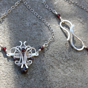 May include: A silver necklace with a pendant of wire-wrapped design and a teardrop gemstone. The necklace has small, round, red gemstones and a chain with a hook clasp. The necklace is made of silver.