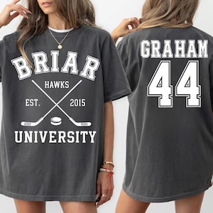 Könnte beinhalten: Ein anthrazitfarbenes Kurzarm-T-Shirt mit weißem Siebdruck. Die Vorderseite zeigt "BRIAR HAWKS UNIVERSITY EST. 2015" mit gekreuzten Hockeyschlägern und einem Puck. Die Rückseite zeigt den Namen "GRAHAM" über der Nummer "44".
