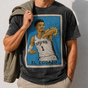 Op de afbeelding: Donkergrijs t-shirt met een illustratie van een basketballer in een blauwe lijst. De speler draagt een Spurs-shirt met nummer 1. De tekst "EL CODAZO" staat onderaan de lijst.