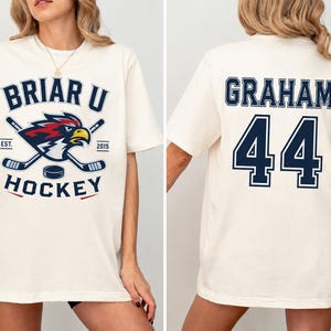 Può includere: Una t-shirt a maniche corte color bianco sporco con una grafica Briar U Hockey sul davanti che raffigura una mascotte falco, bastoni da hockey e un disco. Il retro della maglia mostra il nome Graham e il numero 44 in grandi lettere blu scuro.