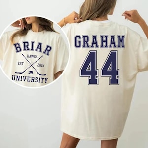 Può includere: Una t-shirt color bianco sporco con testo e grafiche blu scuro. Il retro mostra il nome GRAHAM e il numero 44. L'inserto circolare anteriore mostra il testo BRIAR HAWKS EST. 2015 UNIVERSITY con bastoni da hockey incrociati e un disco.